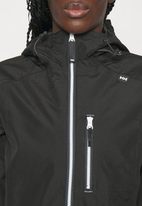 Femme portant une veste noire avec une fermeture éclair blanche et une poche poitrine, avec un petit logo HH sur la poitrine et des cordons de capuche.