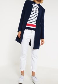 En person med en marinblå kavaj över en vit, marinblå och röd randig polo, vita uppvikta byxor och vita sneakers som står mot en vit bakgrund.
