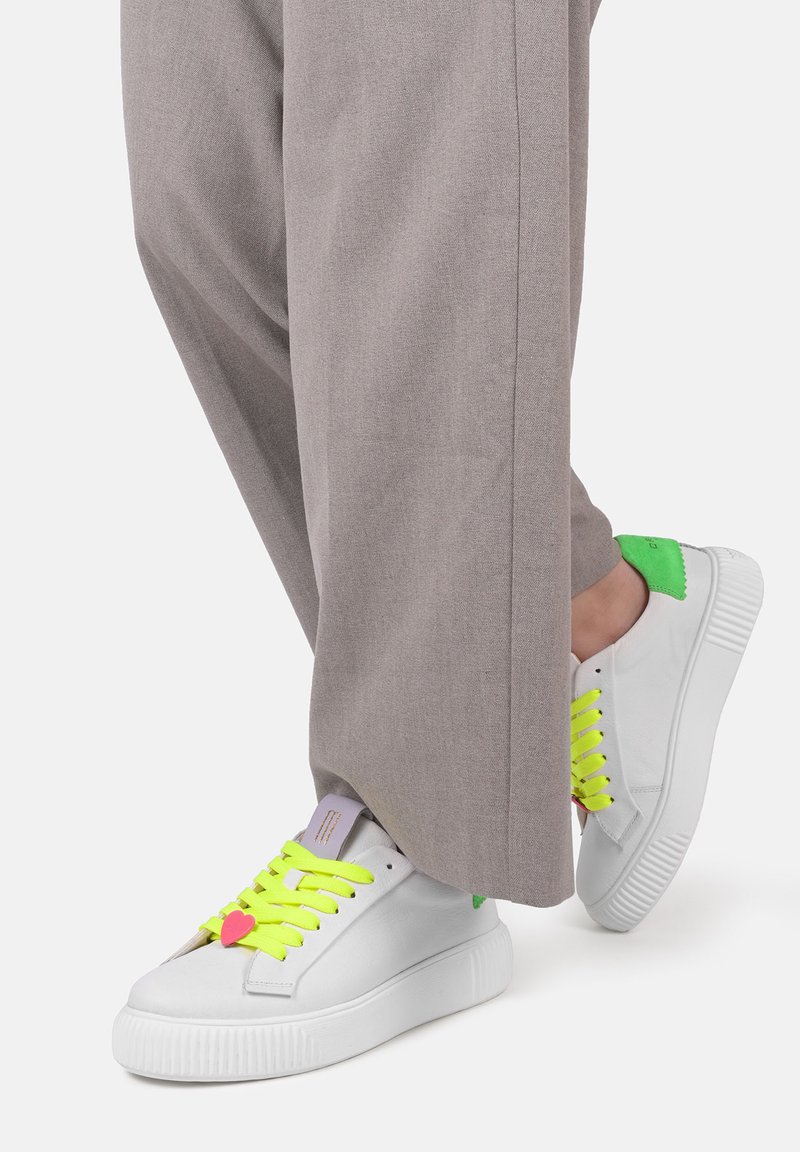 Sneaker bianche con vivaci lacci gialli, dettagli verde sul tallone e una finitura texturizzata. Abbinati a pantaloni grigi larghi.