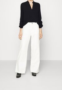 Blusa nera a maniche lunghe con scollatura a V, abbinata a pantaloni bianchi a vita alta e gamba larga. Stivali neri alla caviglia completano il look.