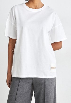 Persoon draagt een los wit T-shirt met korte mouwen en een klein beige label op de zoom en een grijze geplooide broek.