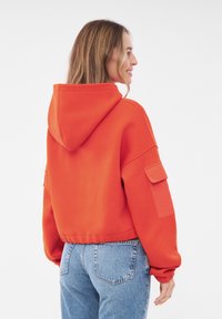 Oranje cropped hoodie met een capuchon en zijzakken, gemaakt van zachte stof. Heeft ribgebreide manchetten en een relaxed fit, gecombineerd met blauwe jeans.