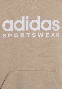 Beige huvtröja i mjukt tyg med en stor vit "adidas"-logotyp och ordet "SPORTSWEAR" nedanför. Den har en framficka och texturerad sömnad.