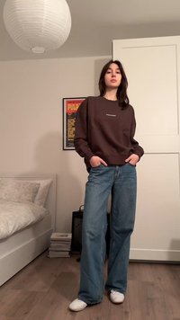 Καφέ φούτερ με άσπρα γράμματα, συνδυασμένο με ανοιχτό μπλε τζιν παντελόνι με φαρδιά μπατζάκια. Το μοντέλο στέκεται με τα χέρια στις τσέπες πάνω σε ξύλινο πάτωμα.