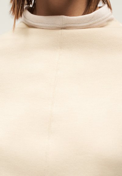 Krämfärgad ribbad tröja med hög krage, med ett texturerat tyg och synliga sömmar längs sömmarna.