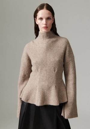 Un mannequin aux cheveux foncés porte un pull beige à col roulé et à côtes, avec un ourlet évasé et des manches longues, associé à une jupe noire sur un fond gris uni.