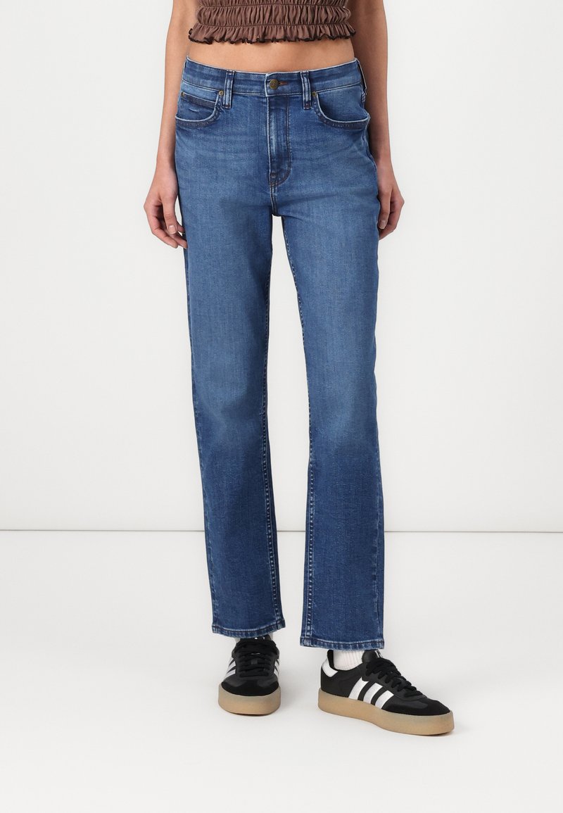 Jean en denim bleu à jambe droite avec une légère décoloration, quatre poches et une fermeture à bouton. Associé à des baskets noires avec des rayures blanches.