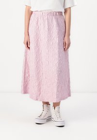 Rosa, teksturert lang skjørt med elastisk midje. Brukt med hvite høye sneakers, som viser en uformell, avslappet passform.