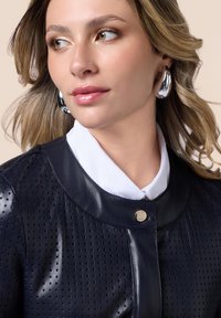 Giacca in pelle perforata blu navy con colletto arrotondato e chiusura a scatto dorata, abbinata a una camicia bianca e orecchini a cerchio argentati.