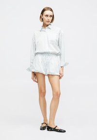 Γυναίκα που φοράει λευκό ριγέ πουκάμισο με μακριά μανίκια και ταιριαστό σορτς, με μαύρα φλατ παπούτσια διακοσμημένα με ασημένιες τρουκς.