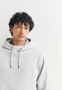 Sweat à capuche gris clair en tissu doux, doté d'une capuche avec cordon de serrage et d'une coupe décontractée. Les détails remarquables incluent des accessoires et des coutures de couleur argentée.