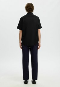 Homme aux cheveux bouclés portant une chemise noire à manches courtes, un pantalon bleu foncé et des chaussures noires, debout de dos sur un fond uni.