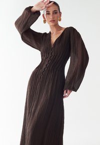 Rjava maksi obleka z nagubami in globokim V-izrezom, dolgimi napihnjenimi rokavi, gumbi po sprednji strani ter teksturirano, tekočo tkanino.