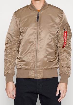 Mand iført beige bomberjakke med lynlås foran, ribstrikkede manchetter og kant samt rød "Remove Before Flight"-mærkat på venstre ærmelomme.