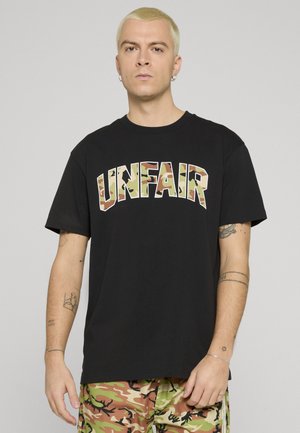 Mann med kort blondt hår iført svart skjorte med "UNFAIR" kamuflasjetekst og kamuflasjemønstrede bukser, står foran en ensfarget lys grå bakgrunn.