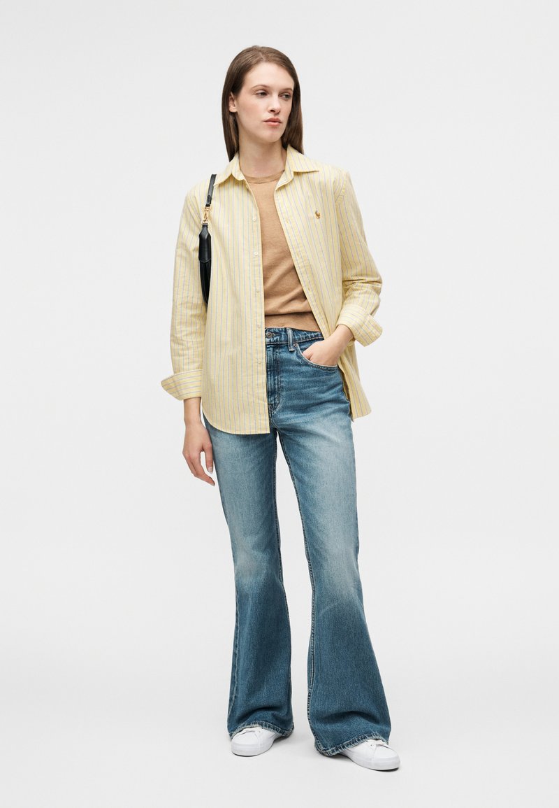 Chemise à rayures jaunes portée sur un haut marron, associée à un jean évasé bleu clair et des baskets blanches. Accent avec un sac à main noir.