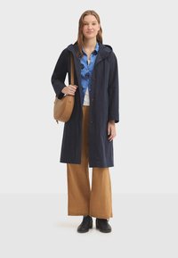 Manteau long marine avec capuche, boutons marron, porté sur une chemise bleue à motifs, un sous-pull crème et un pantalon large beige ; chaussures et sac noirs.