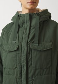 Kvinna som bär en grön vadderad jacka med huva, framdragkedja, två bröstfickor med klaff och långa mörkgröna dragsnören.