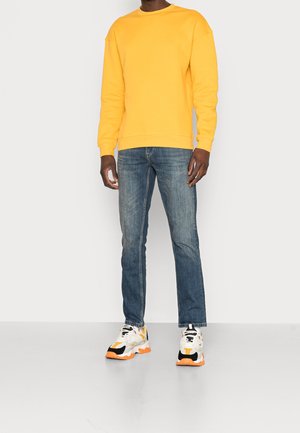 Geel sweatshirt van katoen, gecombineerd met blauwe jeans en witte sneakers met oranje accenten. Eenvoudig, ontspannen ontwerp met ribbels op de mouwen.
