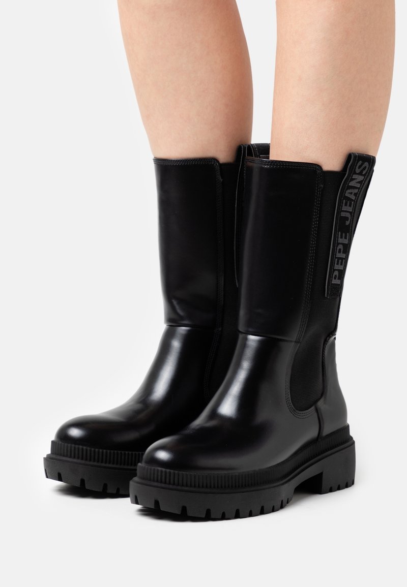 Bottines noires à finition brillante, avec des panneaux latéraux élastiques et des semelles en caoutchouc texturé. Présentent un marquage "PEPE JEANS" sur le côté.