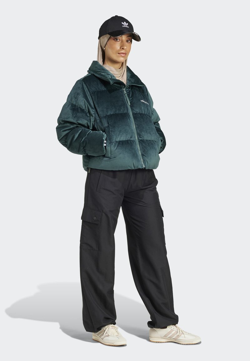 Grüne Samt-Puffermantel mit Reißverschluss und Patches, kombiniert mit schwarzen Cargo-Hosen und cremefarbenen Sneakers; Modell trägt eine schwarze Kappe.