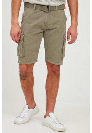 Homme portant un short cargo vert olive avec des poches latérales, une ceinture, des baskets blanches et un tatouage visible sur la jambe, se tenant sur un fond blanc.