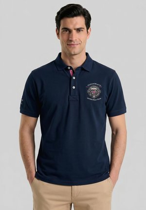 Hombre con una camiseta polo azul marino con el logo de United Polo Team y pantalones caqui, manos en los bolsillos, de pie frente a un fondo gris liso.