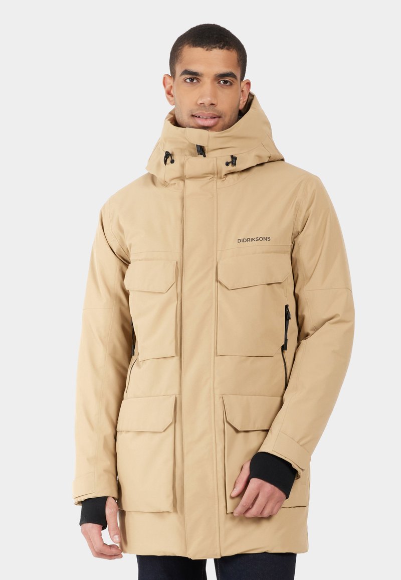 Beige geïsoleerde jas met een hoge kraag, meerdere voorzakken, verstelbare capuchon en zwarte manchetten. Materiaal lijkt glad en duurzaam.