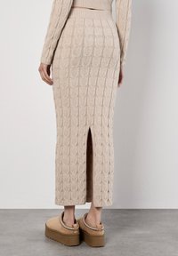 Beige gestrickter Midirock mit strukturiertem Zopfmuster und einem Schlitz hinten. Kombiniert mit beigefarbenen Plateau-Schlupfschuhen für zusätzliche Details.