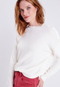 Maglione bianco a coste con scollo rotondo e bottoni decorativi sulla spalla, abbinato a pantaloni rossi. Texture liscia, vestibilità rilassata.