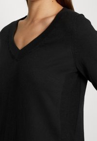 Femme portant un pull en maille noir à col en V, montrant l'encolure, l'épaule et le haut de la poitrine sur un fond uni.