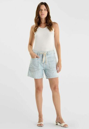 Vrouw draagt wit mouwloos topje en lichtblauwe gestreepte korte broek met trekkoord, staat met één hand in de zak tegen een effen achtergrond.