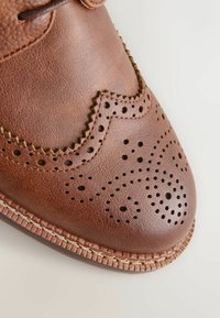 Chaussure brogue en cuir marron avec des perforations décoratives et une couture détaillée sur le bout et les bords.