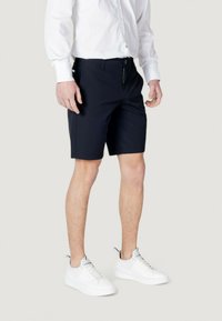 Pantaloni corti blu navy realizzati in tessuto leggero, con chiusura a zip e bottone, indossati con una camicia bianca e sneaker bianche.