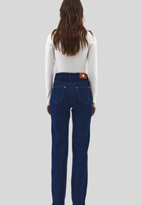 Žena s dlouhými hnědými vlasy, oblečená v bílém topu s dlouhým rukávem a tmavě modrých džínách s vysokým pasem, stojící na jednobarevném světle šedém pozadí.