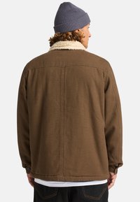 Brown canvas jacket with a cream sherpa collar, button cuffs, and a relaxed fit. Worn with a gray knit beanie.