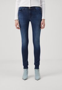 Jeans ajustados en denim azul oscuro con un ligero efecto desgastado, diseño de cinco bolsillos y una cintura de altura media, combinados con botas de tobillo azul claro.