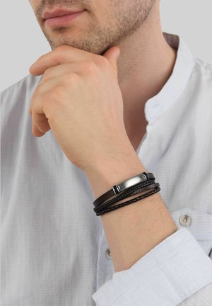 Hombre con camisa blanca apoyando la barbilla en la mano, mostrando una pulsera de cuero trenzado marrón oscuro con cierre metálico en la muñeca.