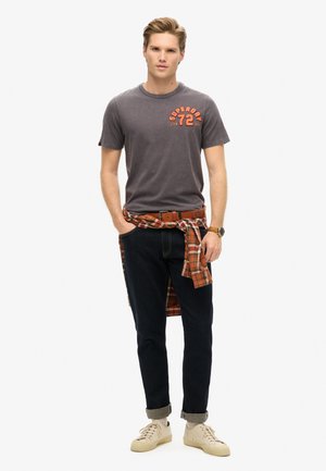 Graues Kurzarm-T-Shirt mit orangem Druck, dunkelblaue Jeans, beiges Sneaker und ein rotkariertes Hemd um die Taille gebunden.