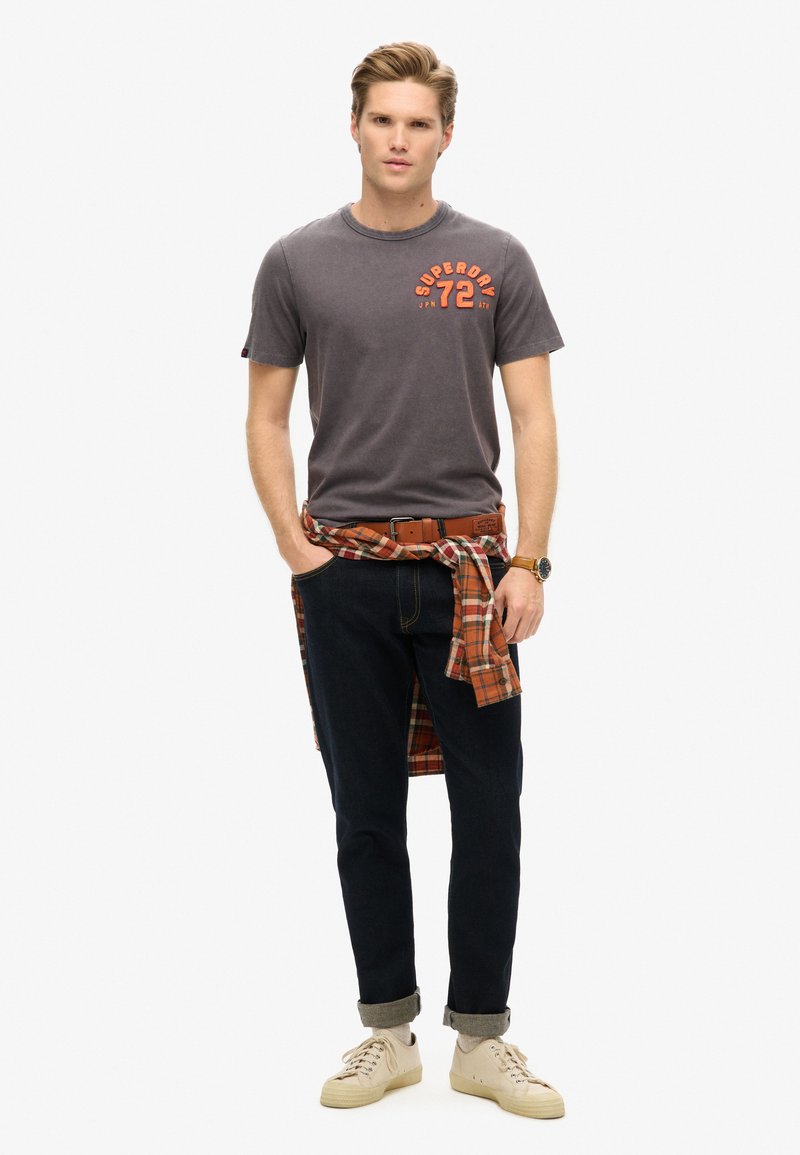 Camiseta gris de manga corta con estampado naranja, pantalones vaqueros azul oscuro, zapatillas beige y una camisa a cuadros roja atada a la cintura.