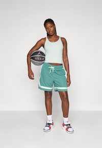 Top deportivo de tirantes verde claro con un logo, combinado con pantalones cortos verdes que tienen rayas blancas y un cordón. Zapatillas negras y blancas.