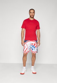 Camiseta roja de manga corta con un logo negro, combinada con pantalones cortos blancos que tienen gráficos de los Yankees en azul y rojo. Zapatillas blancas completan el look.