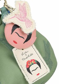 Groene stoffen tas met twee decoratieve labels: een roze rond label met een gezichtontwerp en een crèmekleurig label met een bloemmotief en handtekening.