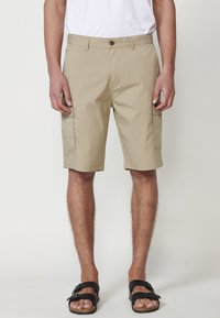 Pantaloni scurți cargo bej cu două buzunare laterale, închidere cu nasture și bucle pentru curea. Material texturat cu un croi lejer, purtați cu sandale negre.