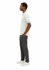 Weißes Poloshirt mit blauen Streifen an den Ärmelabschlüssen, kombiniert mit dunkelgrauen Hosen und weißen Sneakern. Glattes Material, lässige Passform, Seitenansicht.
