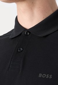 Gros plan d'une personne portant un polo noir avec deux boutons et un petit logo "BOSS" sur la poitrine.