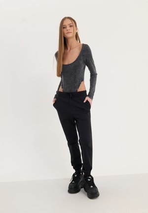 Černý body s dlouhými rukávy a výřezovými boky, spárovaný s černými joggery. Modelka má na sobě masivní černé sneakers. Minimalistické pozadí.