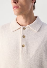 Close-up van een persoon die een gestructureerde off-white gekraagde shirt draagt met drie beige knopen aan de voorkant.