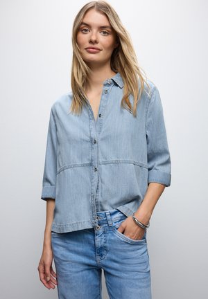 Camisa de mezclilla azul claro con cuello abotonado, botones frontales y mangas remangadas; combinada con unos jeans de mezclilla azul claro a juego.