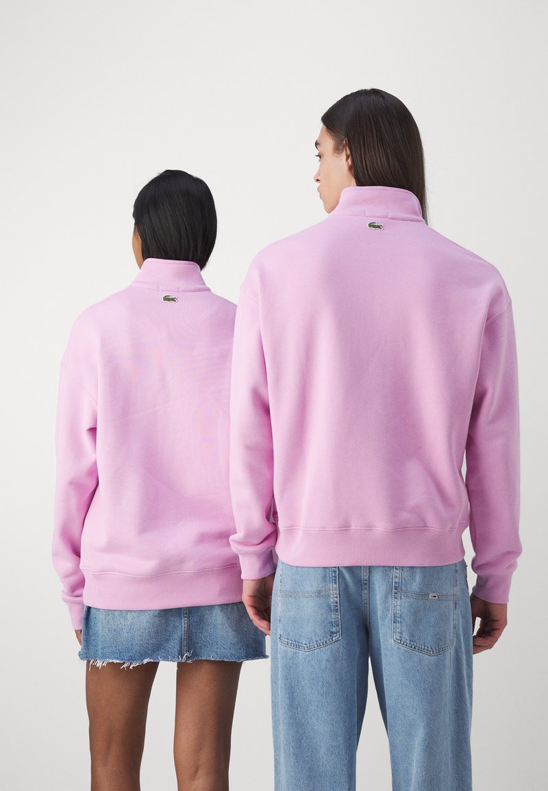 Felpa rosa con colletto alto, polsini e orlo a coste, caratterizzata da un piccolo logo verde sulla parte posteriore. Indossata con pantaloni corti di jeans e jeans chiari.